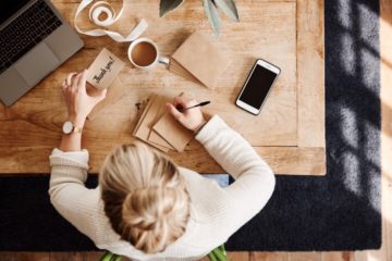 Une photo prise d'en haut d'une femme aux cheveux blonds assise à un grand bureau en bois écrivant à la main "Merci!" sur une pile de petites enveloppes.