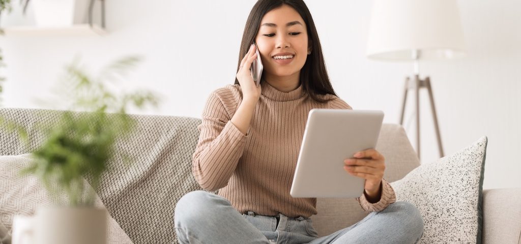 Une jeune femme est assise en tailleur sur un canapé, souriante alors qu'elle regarde un grand bloc-notes et parle sur son téléphone portable.